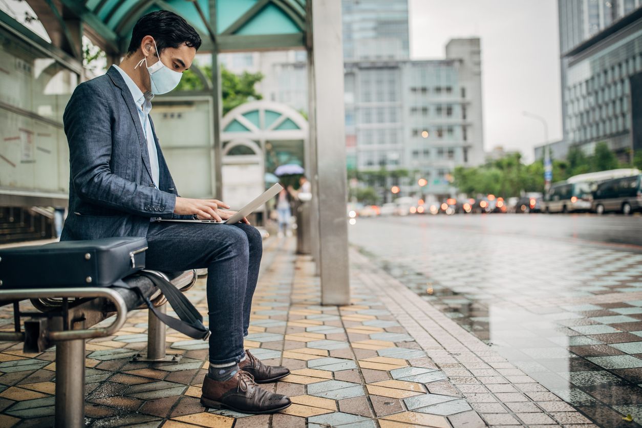 A man in a casual suit sits alone at a bus stop while working at his laptop. The sky is grey and roads wet. 