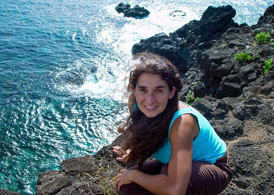 Dr Moira Fortin Cornejo in a blue T-shirt smiling, crouching on a high stone cliff with the ocean below