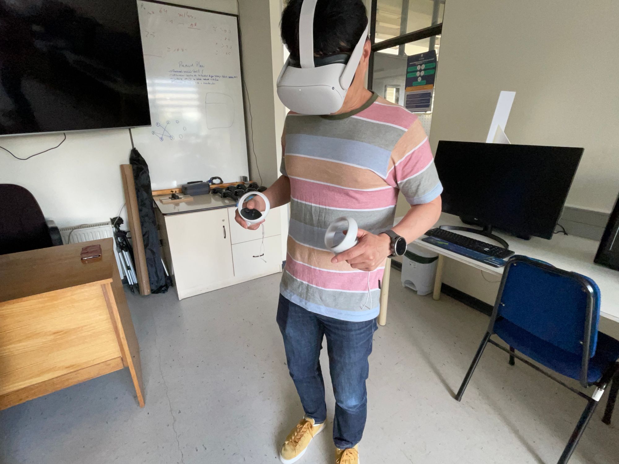 In a small computer lab, a man stands wearing a virtual reality (VR) headset, grasping a VR controller in each hand