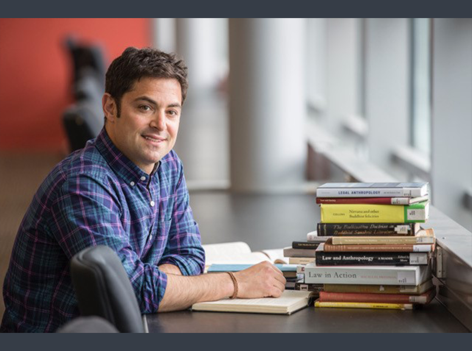 Ben Schonthal at a library table in blue check shirt, with a stack of books 