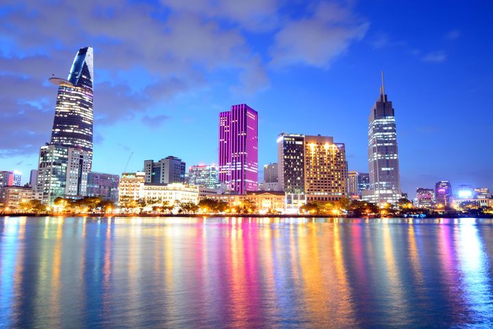 At night, modern office buildings reflect purple and orange lights onto a calm river