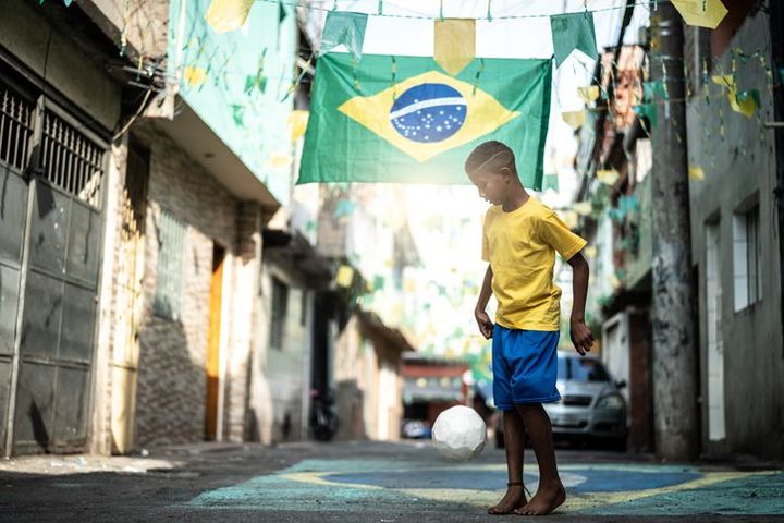 A boy in yellow and blue practicing football in the alleyway of a favela, with the Brazilian flag above him 